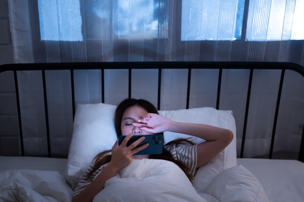 Young Asian woman in bed covering eyes due to glare from smartphone screen at night